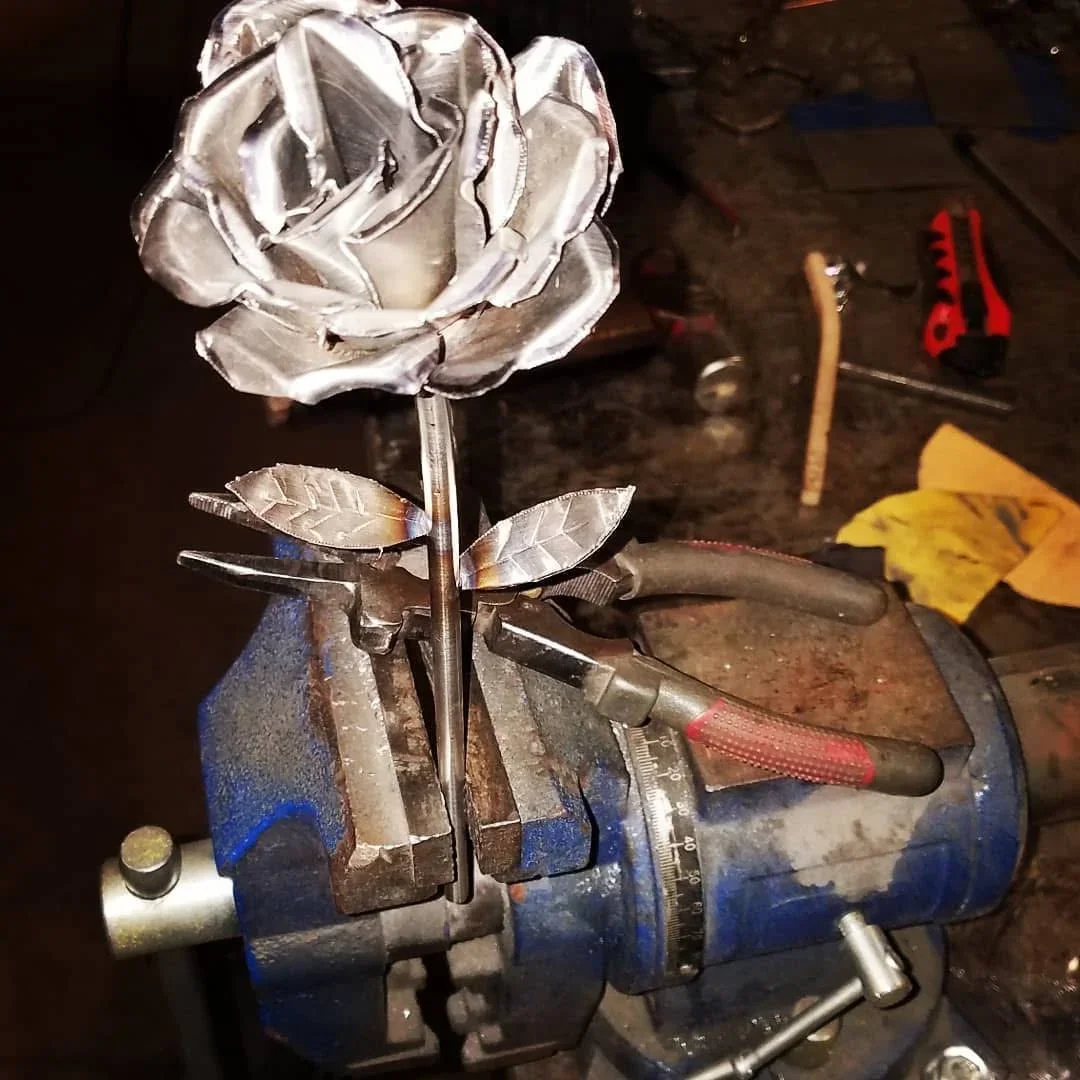 Hand-forged steel rose with detailed petals and leaves held in bench vise during fabrication